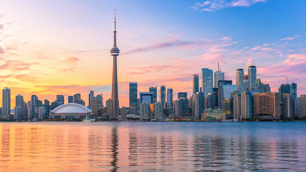 A panoramic photograph of the Toronto city skyline at sunset. The iconic CN Tower and the dome-shaped Rogers Centre stand out prominently among numerous skyscrapers. The sky features vibrant hues of pink, orange, purple, and light blue, and the entire cityscape is beautifully reflected in the calm waters of Lake Ontario in the foreground.